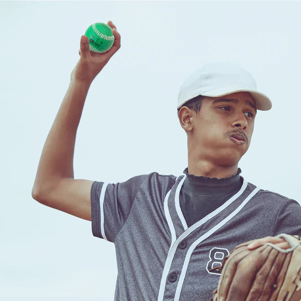 Person in a baseball uniform holding a green Weighted Baseball and a brown glove against a clear sky.