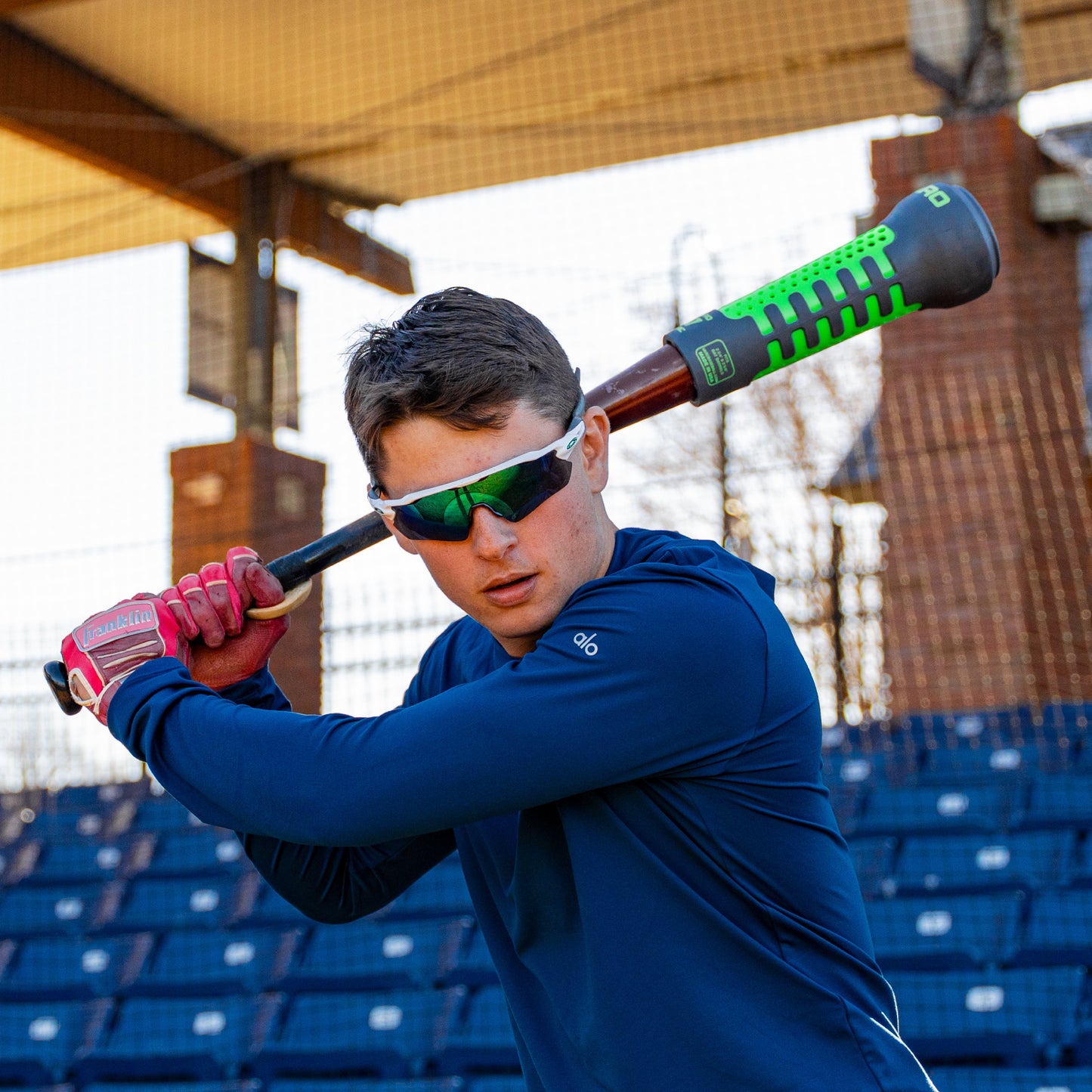 Person in blue shirt and sunglasses holding a baseball bat with Varo ARC Bat Weight 12 Oz on it, in a batting cage.
